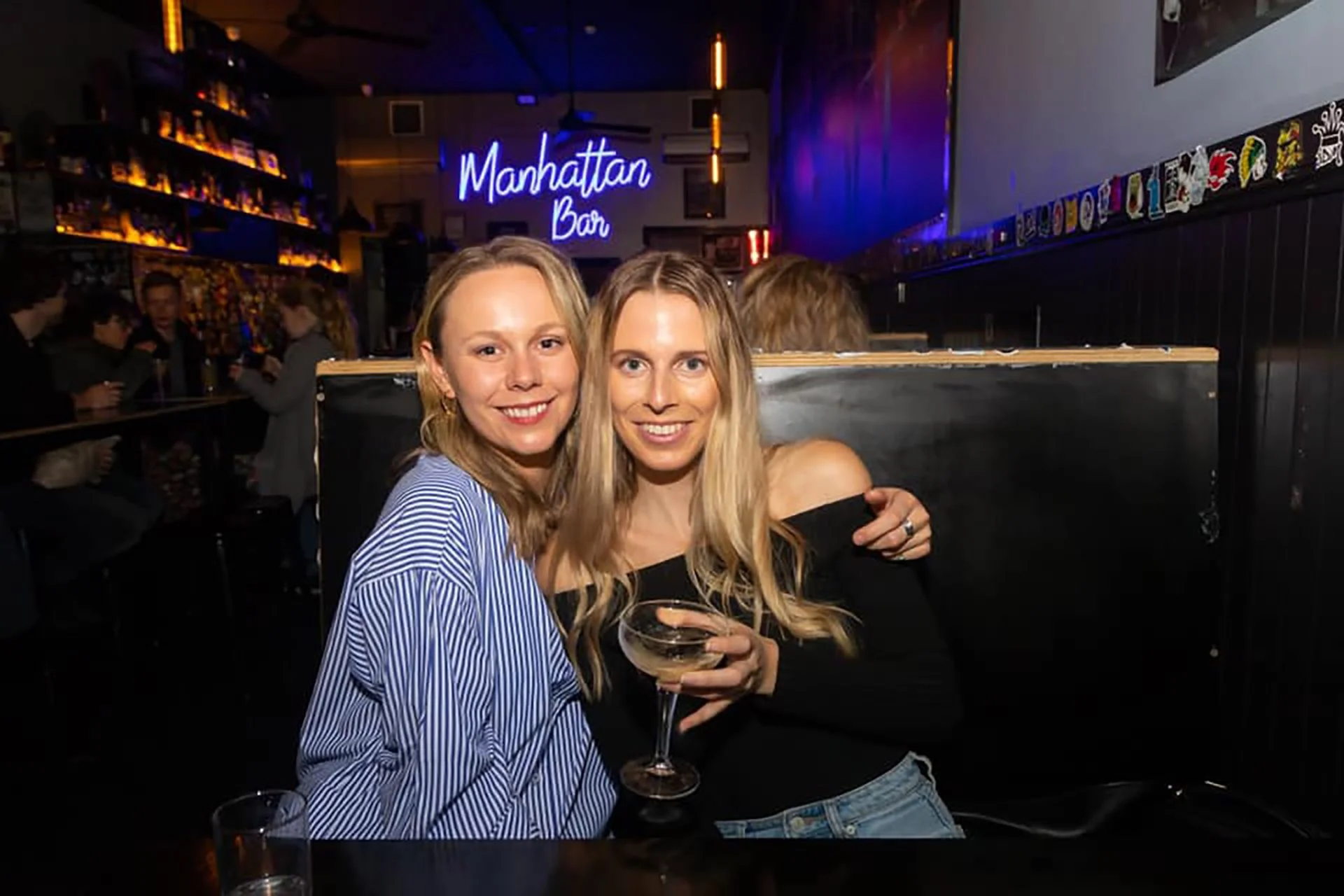 Two women smiling and hugging at a bar with a Manhattan Bar sign in the background, one holding a cocktail glass.