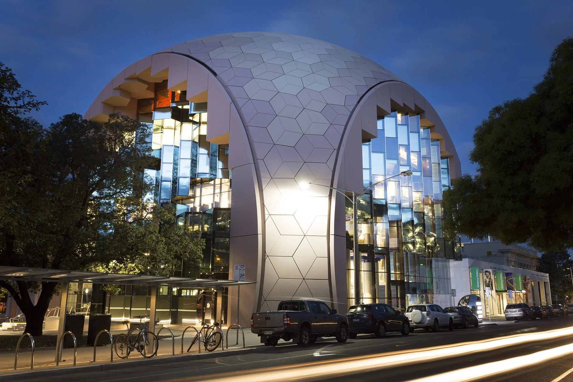 Modern building with a rounded roof and large glass windows, street with parked cars and bicycles, trees, and city lights at night.