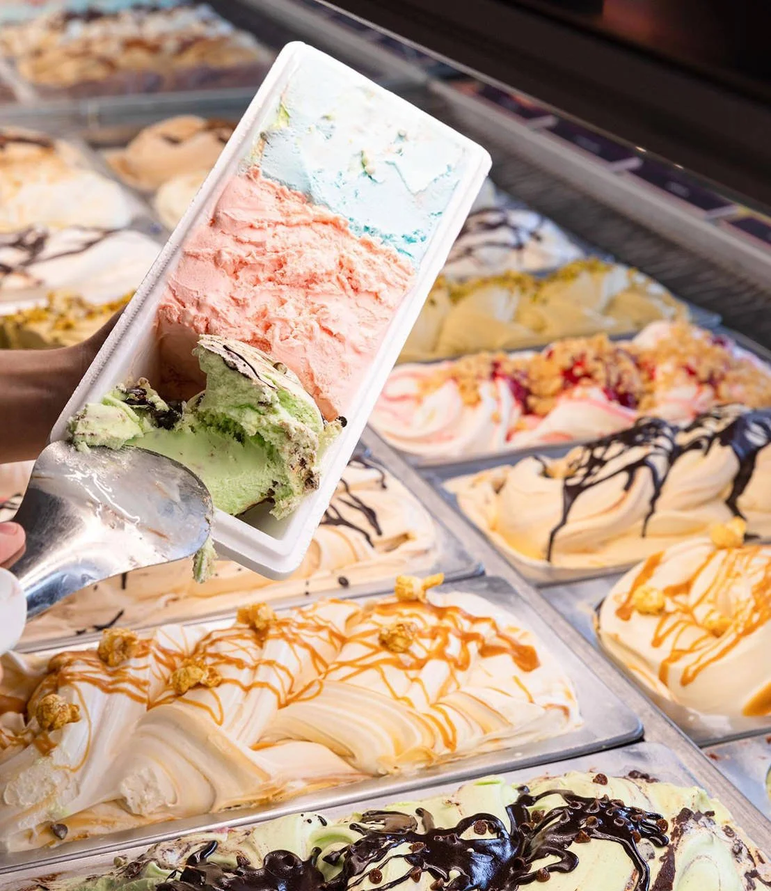 Ice cream shop with various flavors of ice cream in a display case, and a person holding a cup of three-flavor ice cream cone.