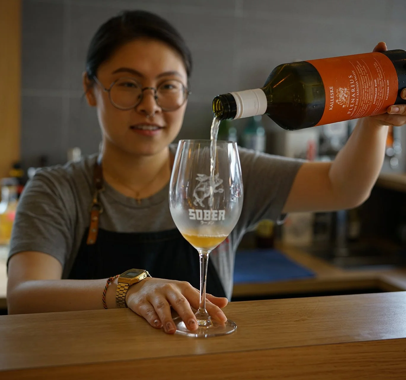 A woman with glasses and a watch pours wine into a glass with the word 'SOBER' and a lion logo, in a kitchen setting.