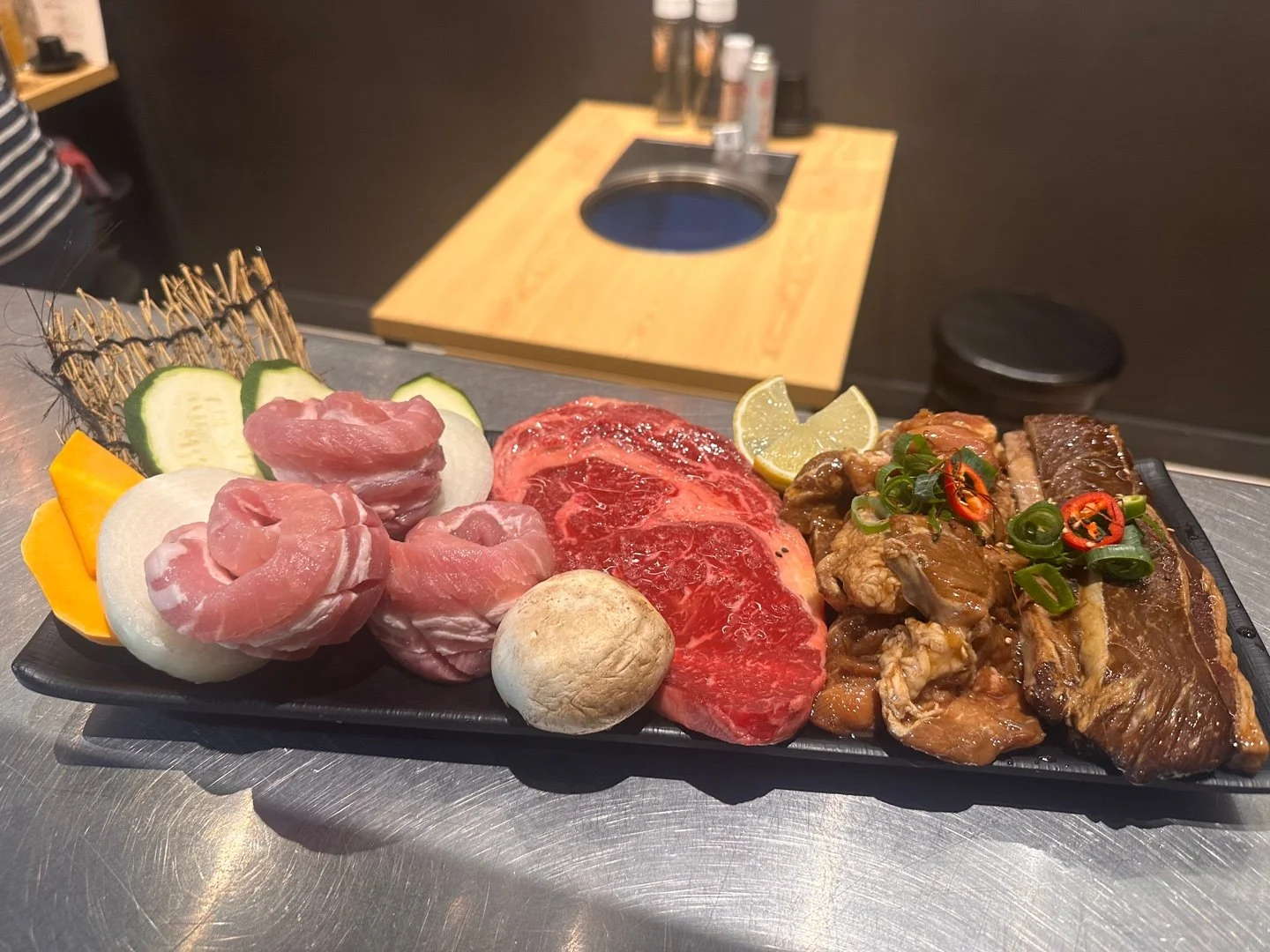 Black rectangular platter with assorted raw meats and vegetables, including slices of cucumber, onion, yellow bell pepper, raw beef, and slices of seasoned beef, garnished with lemon wedges, green onions, and sliced red chili peppers, placed on a sil