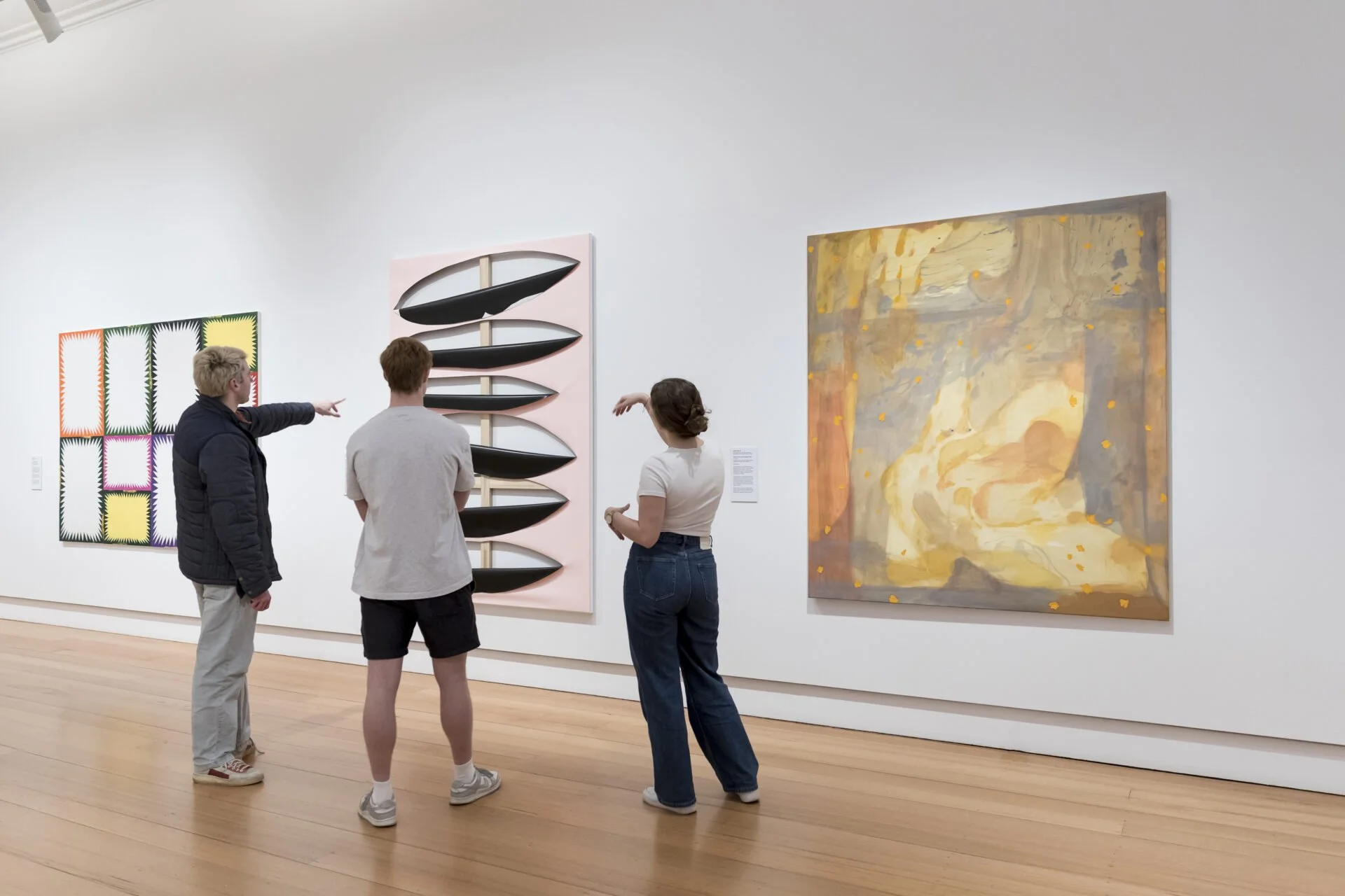 Three people in an art gallery viewed abstract colorful paintings and sculptures on white walls.