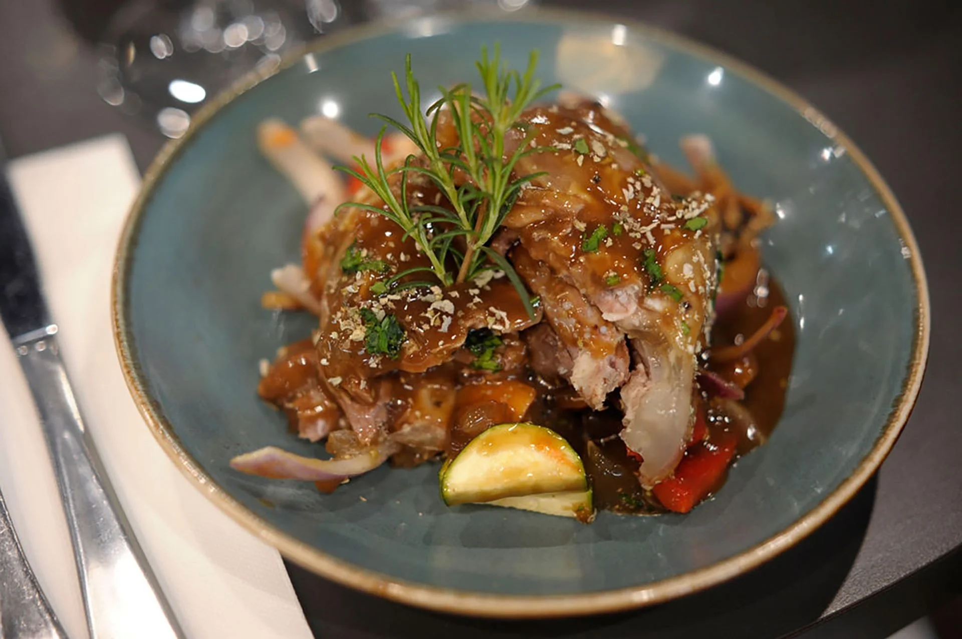 A plated dish featuring a piece of cooked meat topped with a brown sauce, garnished with a sprig of fresh rosemary and chopped herbs, and accompanied by vegetables including a slice of zucchini and others underneath.