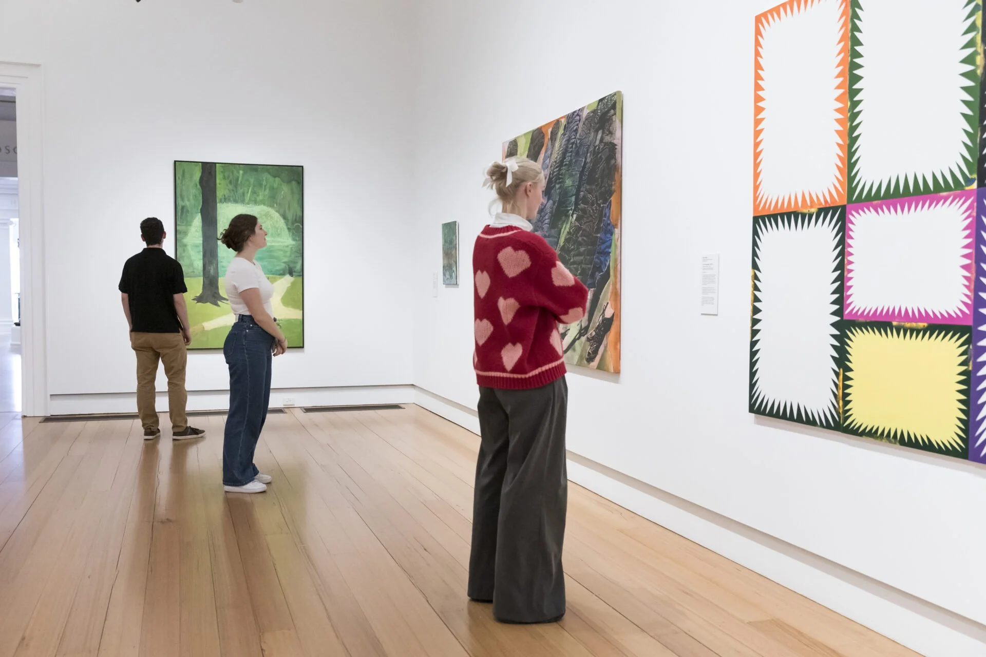 People viewing paintings in an art gallery with white walls and wooden floors.
