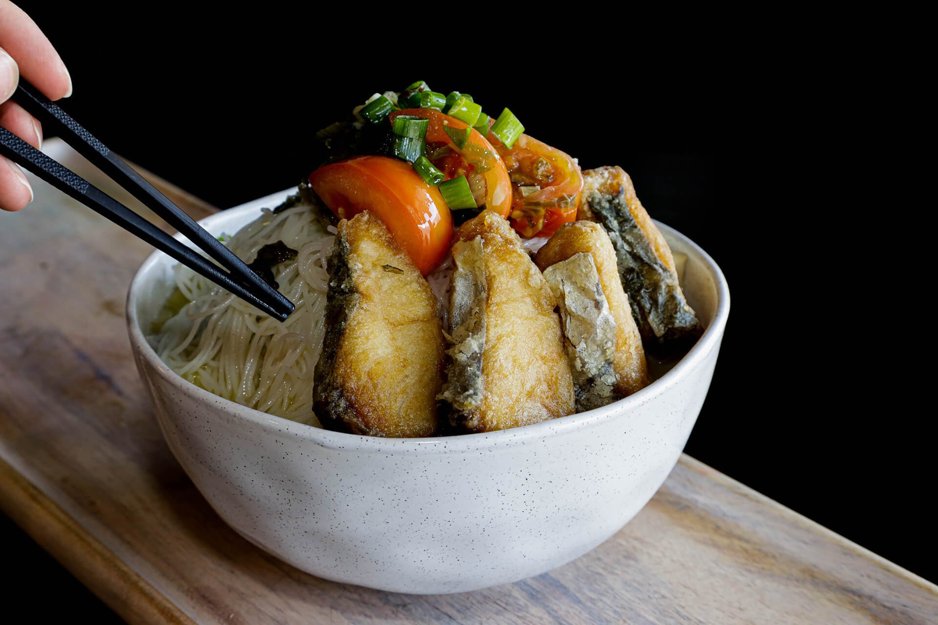 A bowl of Asian noodle soup topped with fried fish fillets, cherry tomatoes, chopped green onions, and vegetables, on a wooden surface.