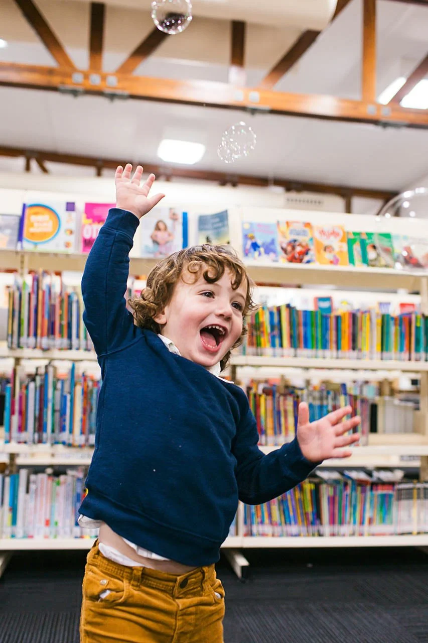 A young boy with curly hair, wearing a navy sweater and mustard-colored pants, joyfully reaching up and smiling in a library with bookshelves filled with colorful books in the background.
