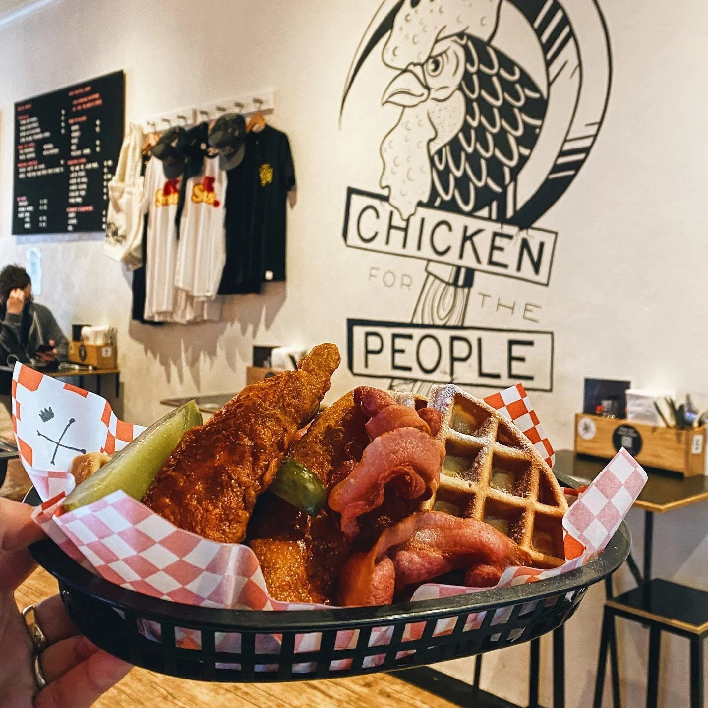 A basket of fried chicken, waffle, bacon, and pickles in a restaurant with a sign on the wall that says 'Chicken for the People' and sports jerseys hanging on the wall.