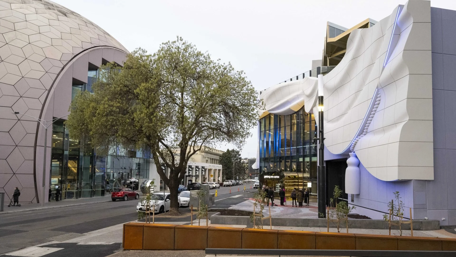 Modern building facades with geometric and organic design elements, a large green tree, parked cars, and pedestrians on the sidewalk.