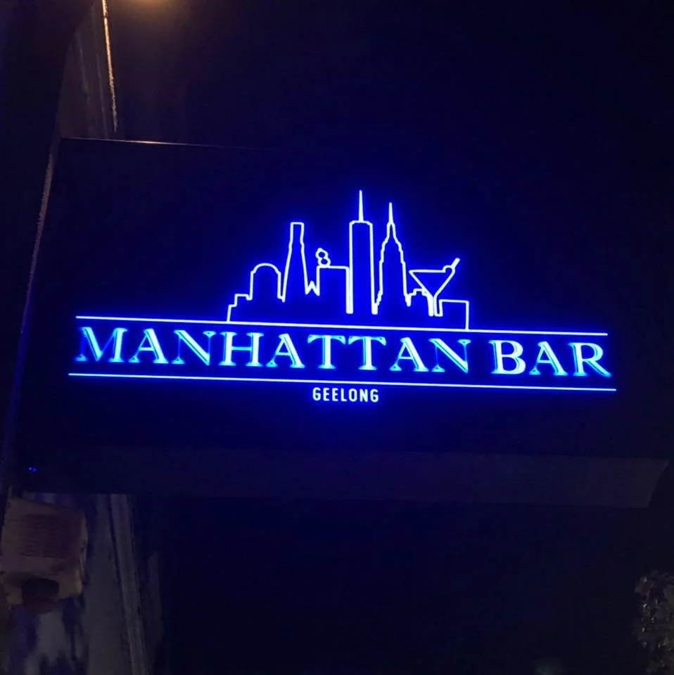 Blue neon sign for Manhattan Bar in Geelong, featuring a stylized skyline of Manhattan with tall buildings and a martini glass.