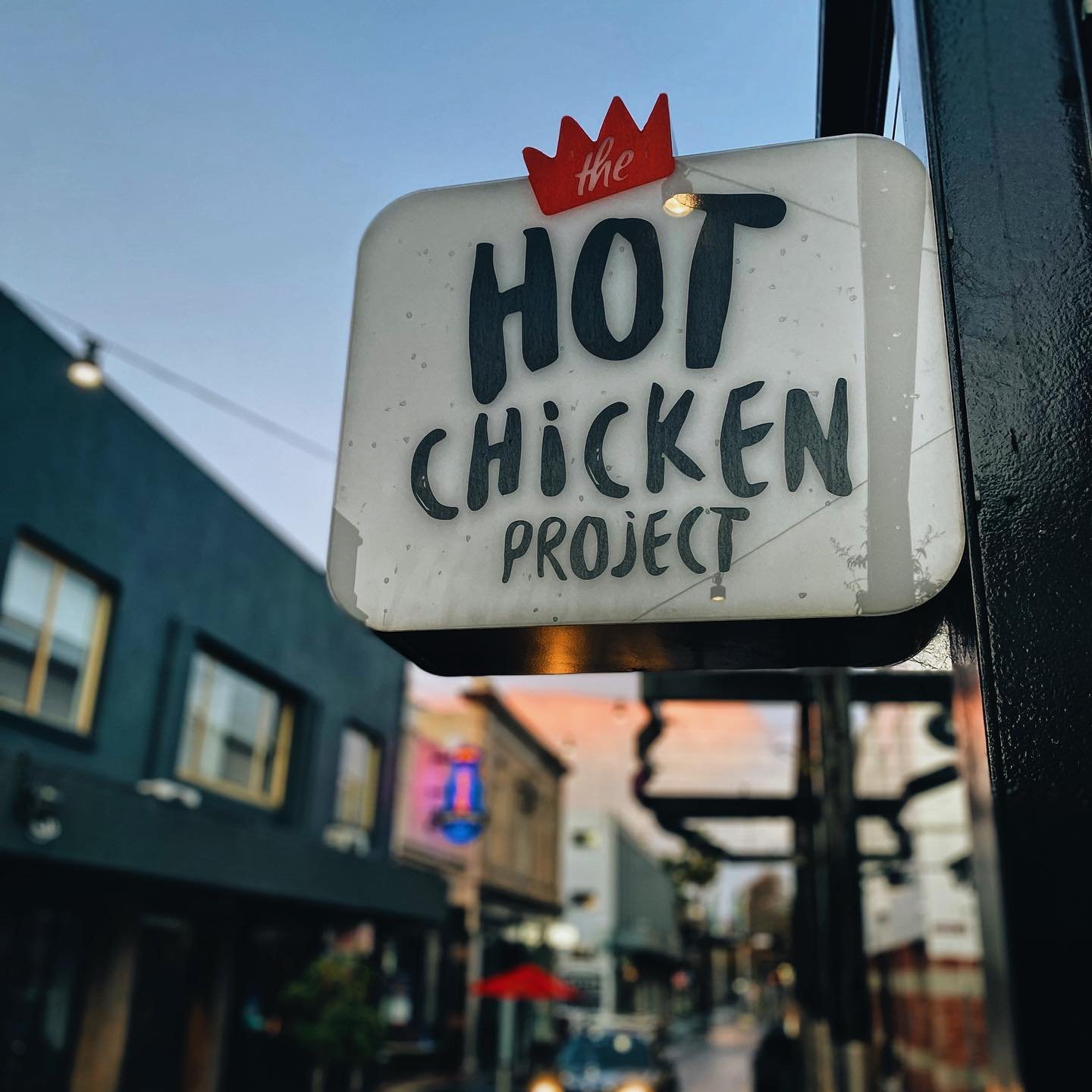 Sign for the Hot Chicken Project hanging outdoors on a building, with a street scene in the background during dusk.