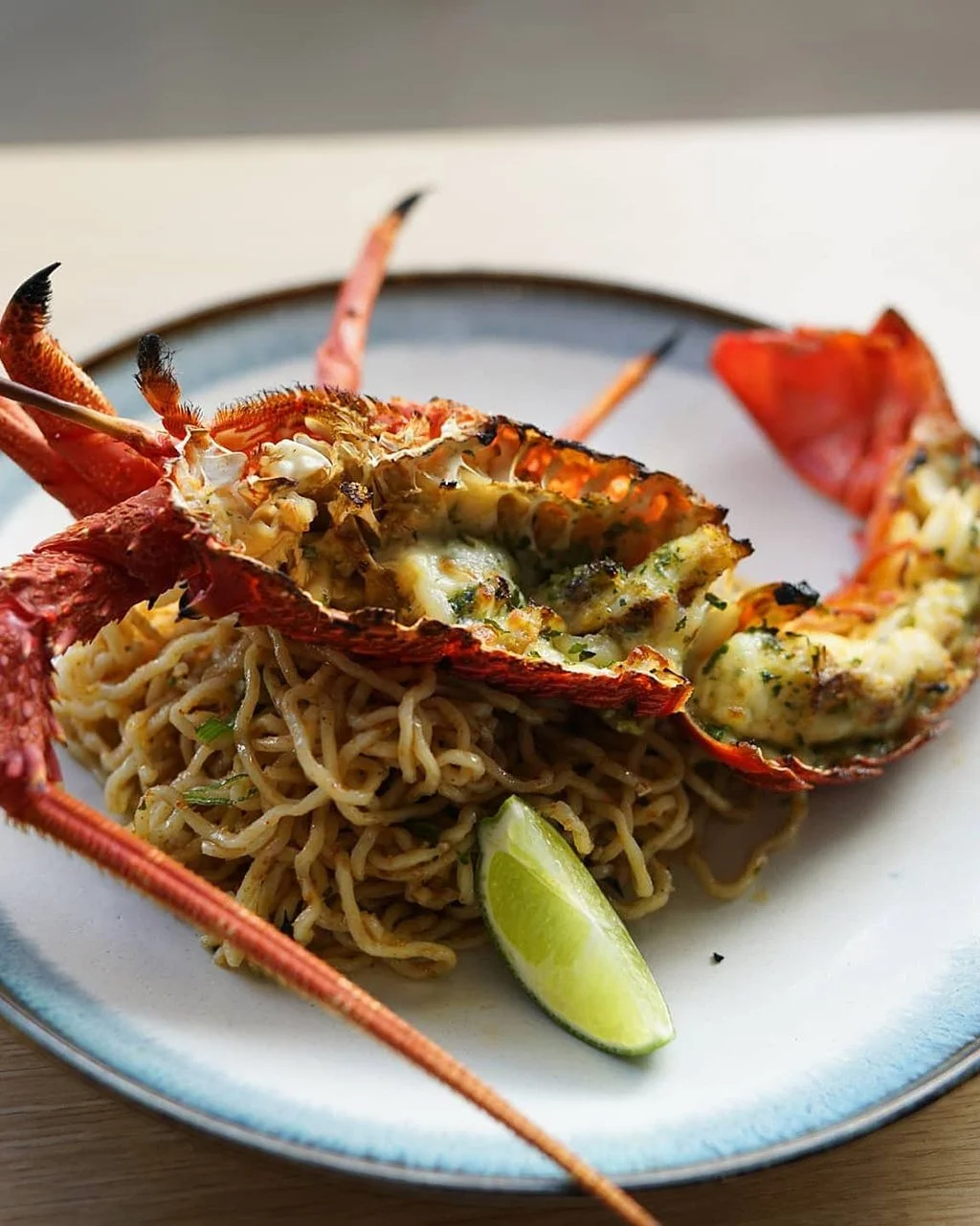 Lobster tail with melted cheese on top, placed over a bed of ramen noodles with chopped green onions, on a white plate with a lime wedge.