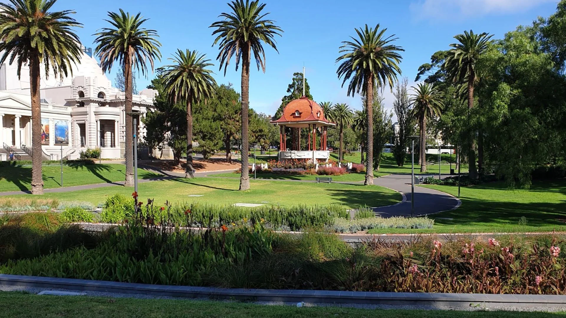 A park with green grass, palm trees, and a red-roofed gazebo in the center. There is a white building with columns and murals in the background under a blue sky.