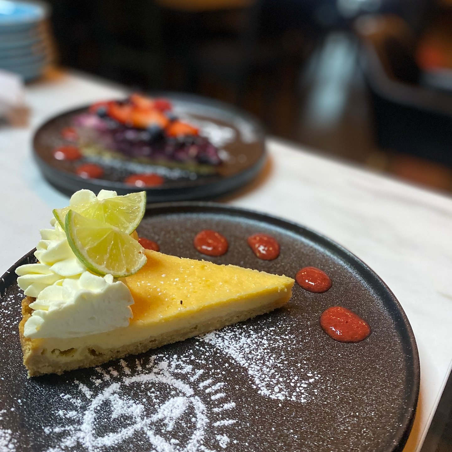 A slice of lemon cheesecake with whipped cream and a lime wedge on top, served on a dark plate with powdered sugar and red sauce dots, with a blurred dessert plate in the background.