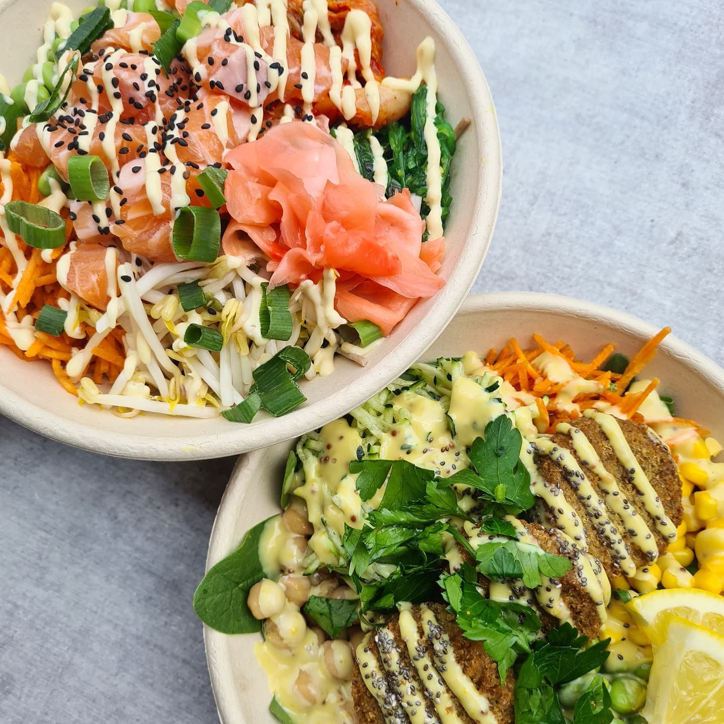 Two bowls of poke bowls with fresh fish, vegetables, sauces, and garnishes including green onions, pickled ginger, avocado, corn, and herbs.