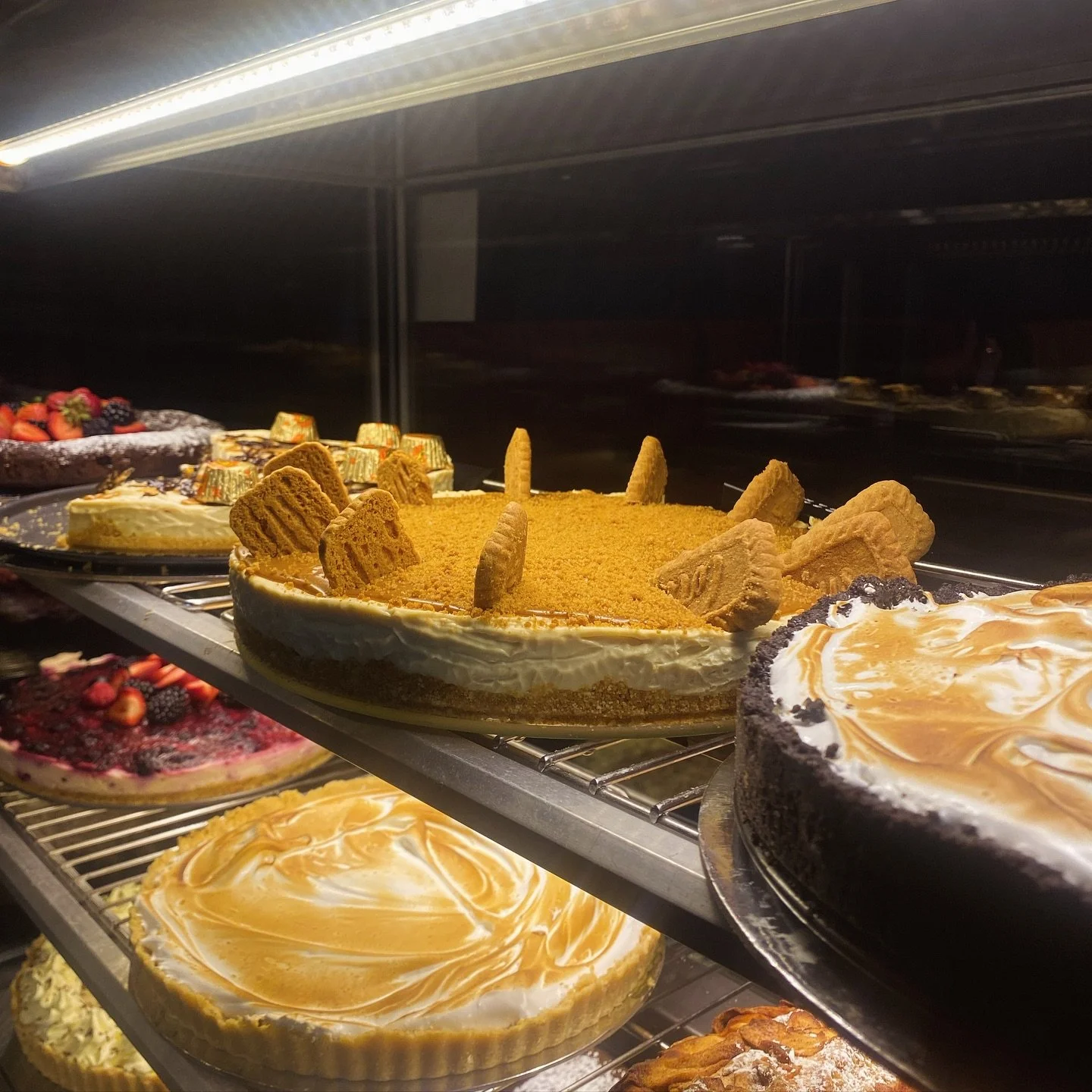 Display of various pies and cakes in a bakery case, including a pie topped with graham crackers, a cheesecake with caramels, and other fruit and cream-topped desserts.