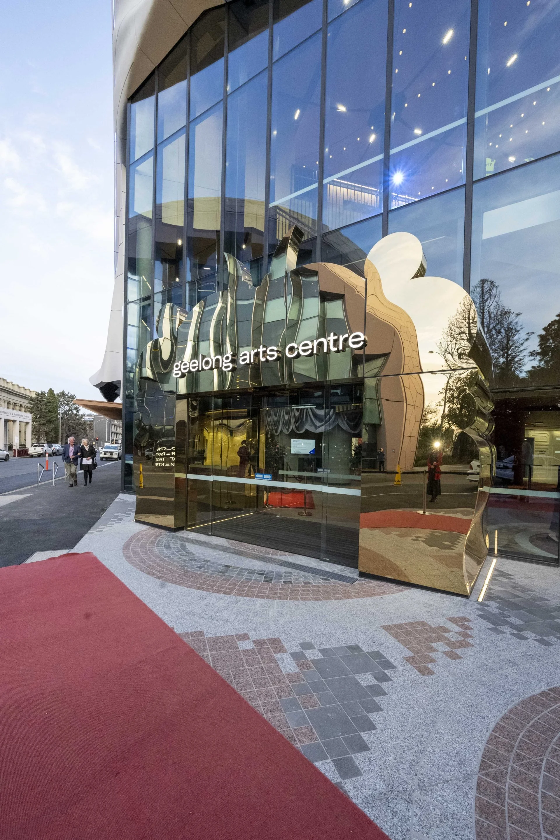Exterior view of the Geelong Arts Centre, a modern building with a curved, glass facade and decorative metallic sculpture above the entrance. There is a red carpet leading to the entrance, and a few people walking nearby.