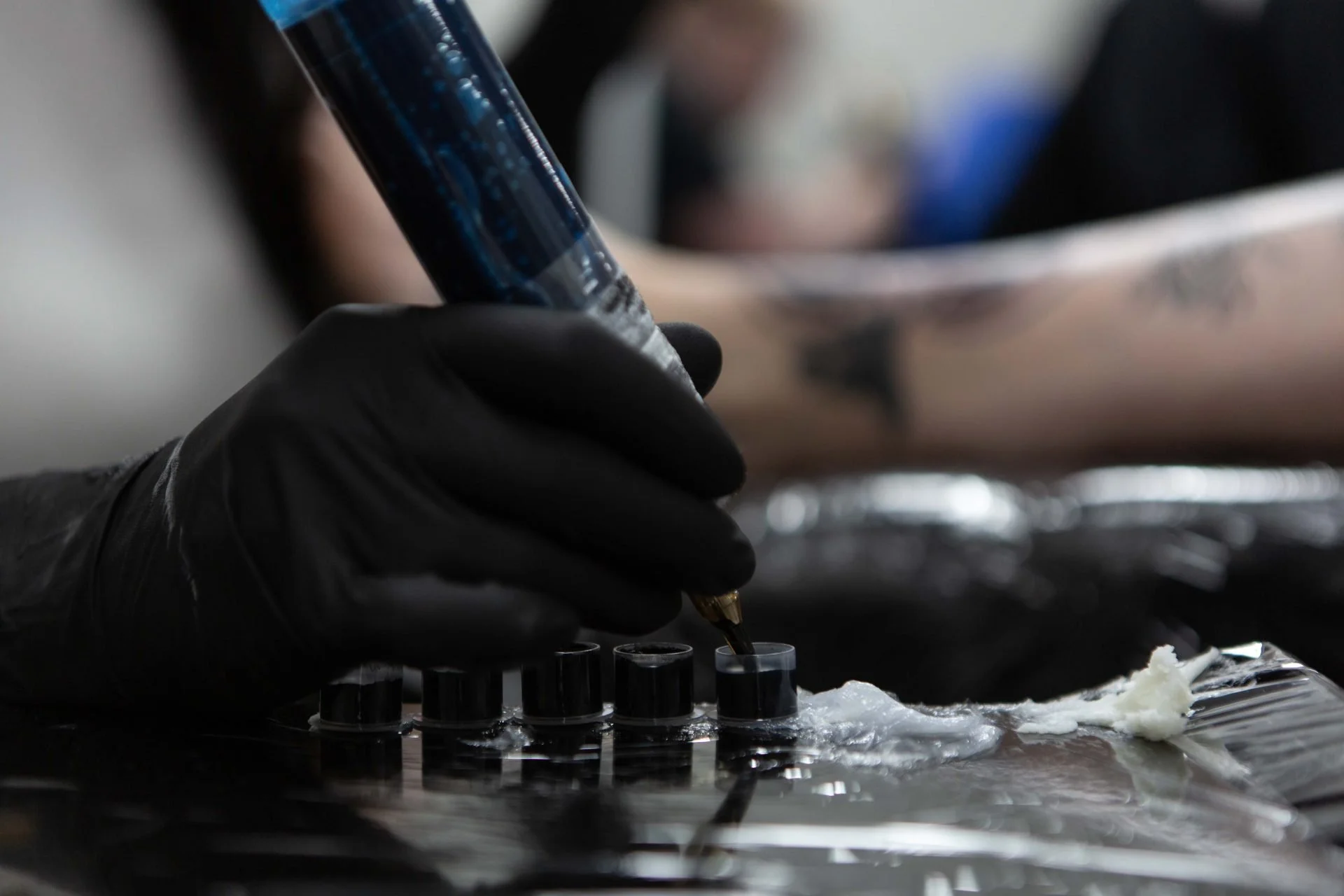 A tattoo artist wearing black gloves uses a tattoo machine to ink a design onto a person's arm, with the arm resting on a surface covered with a plastic sheet.