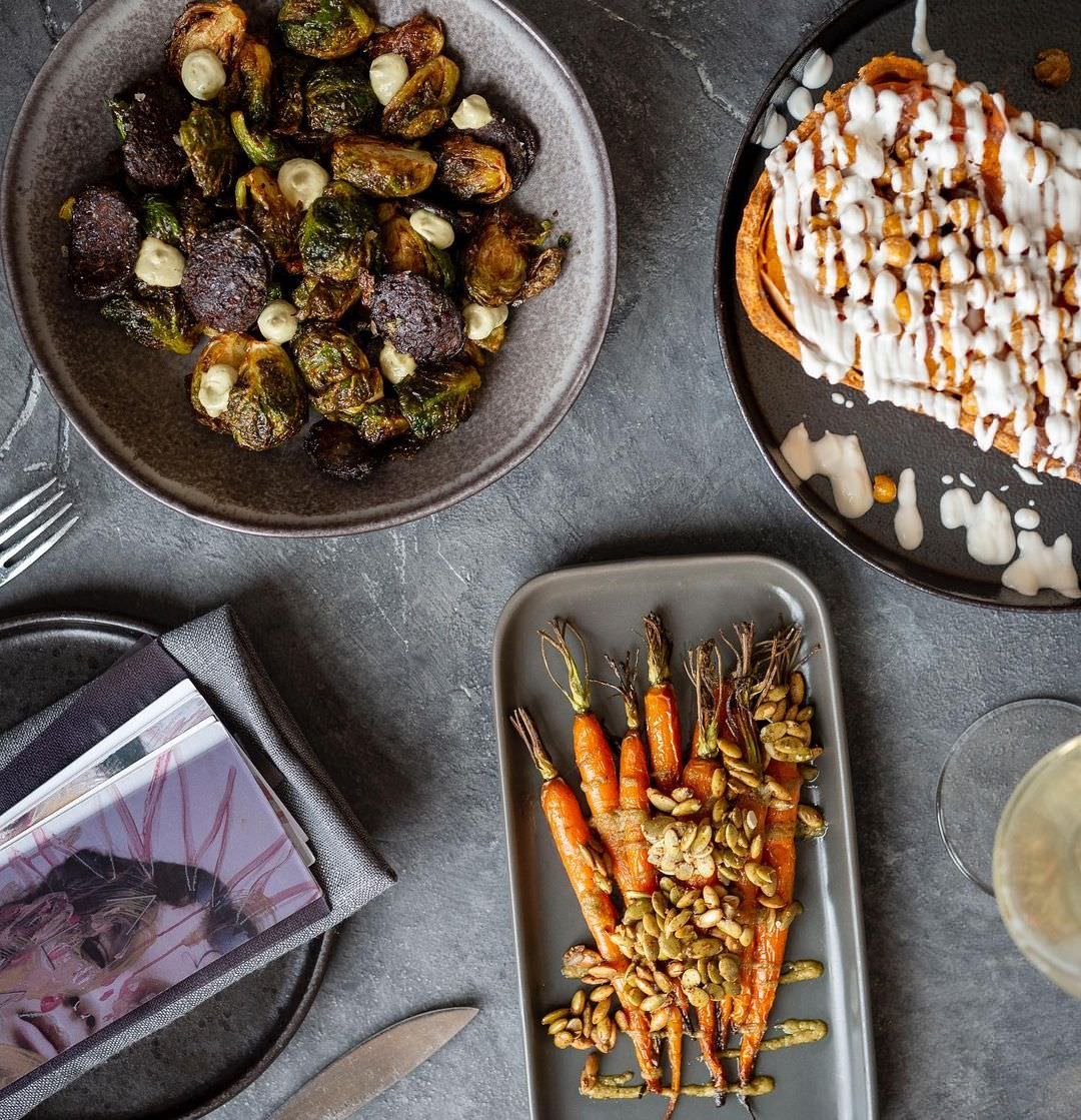 A top-down view of a table with three dishes: a bowl of roasted Brussels sprouts with garlic, a plate of sweet potato casserole with marshmallows and caramel drizzle, and a platter of roasted carrots topped with pumpkin seeds. There is also a wine gl