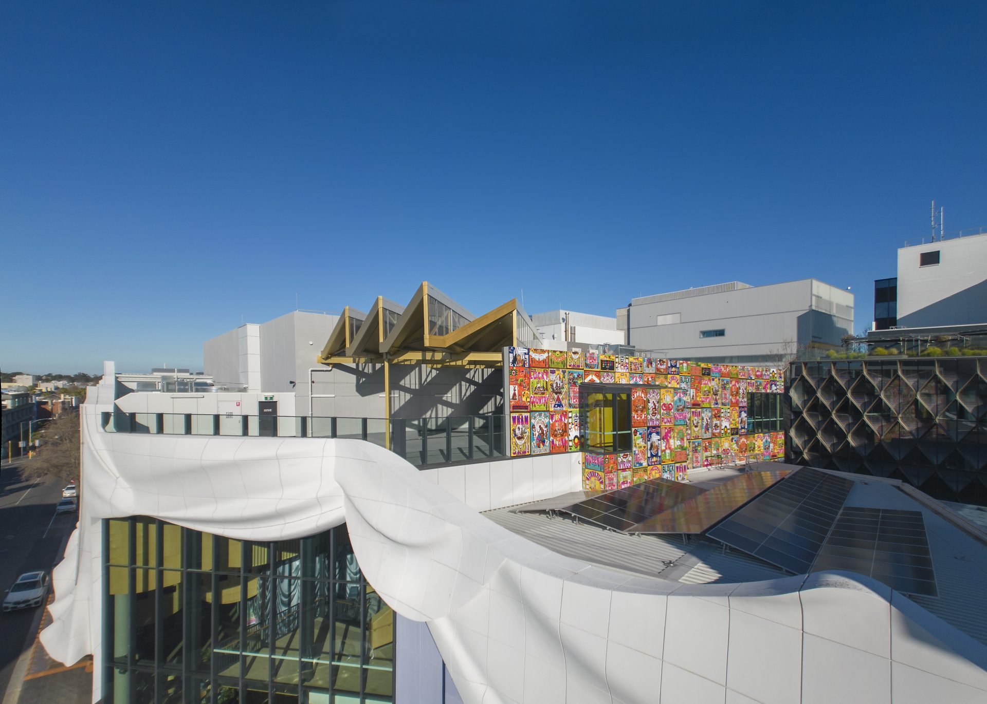 A modern building with white, curved and angular architectural features, including a rooftop mural composed of colorful cartoon and pop art images, against a clear blue sky.