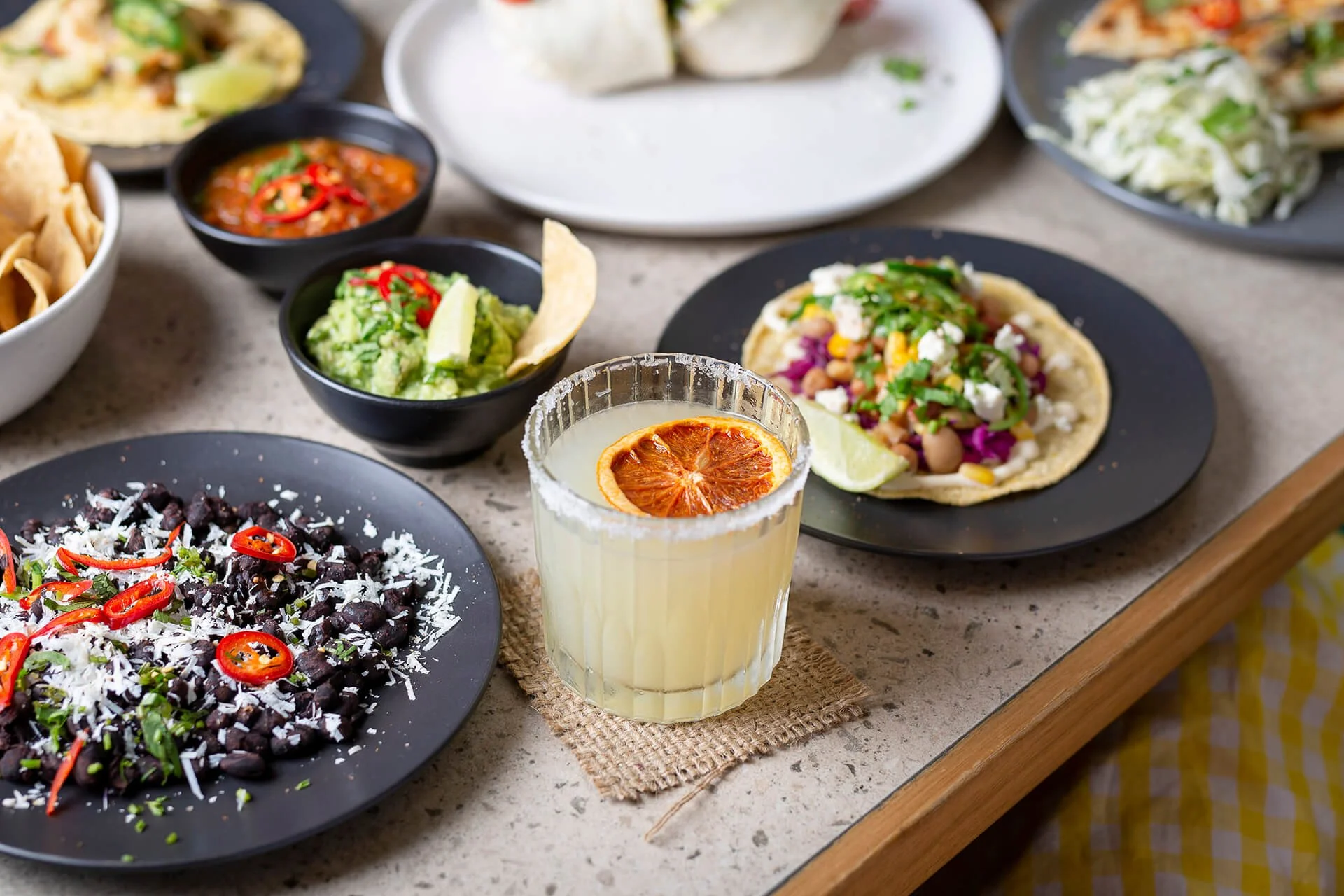 Assorted Mexican dishes on a table, including a spicy black bean salad, guacamole, a margarita with a salted rim and a slice of blood orange, tacos with various toppings, and bowls of salsa and shredded cabbage.