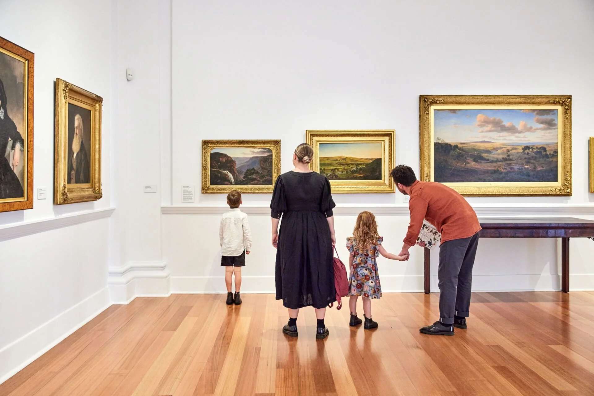 A family of four observing landscape paintings in an art gallery, with a woman, a man, and two children, one boy and one girl. The gallery has white walls and wooden floors.