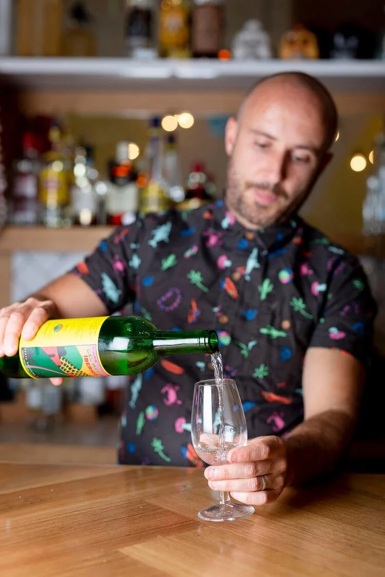 A man pouring a clear beverage into a wine glass in a bar or kitchen setting. He is wearing a black shirt with colorful floral and tropical patterns, and behind him are shelves with bottles and bar supplies.