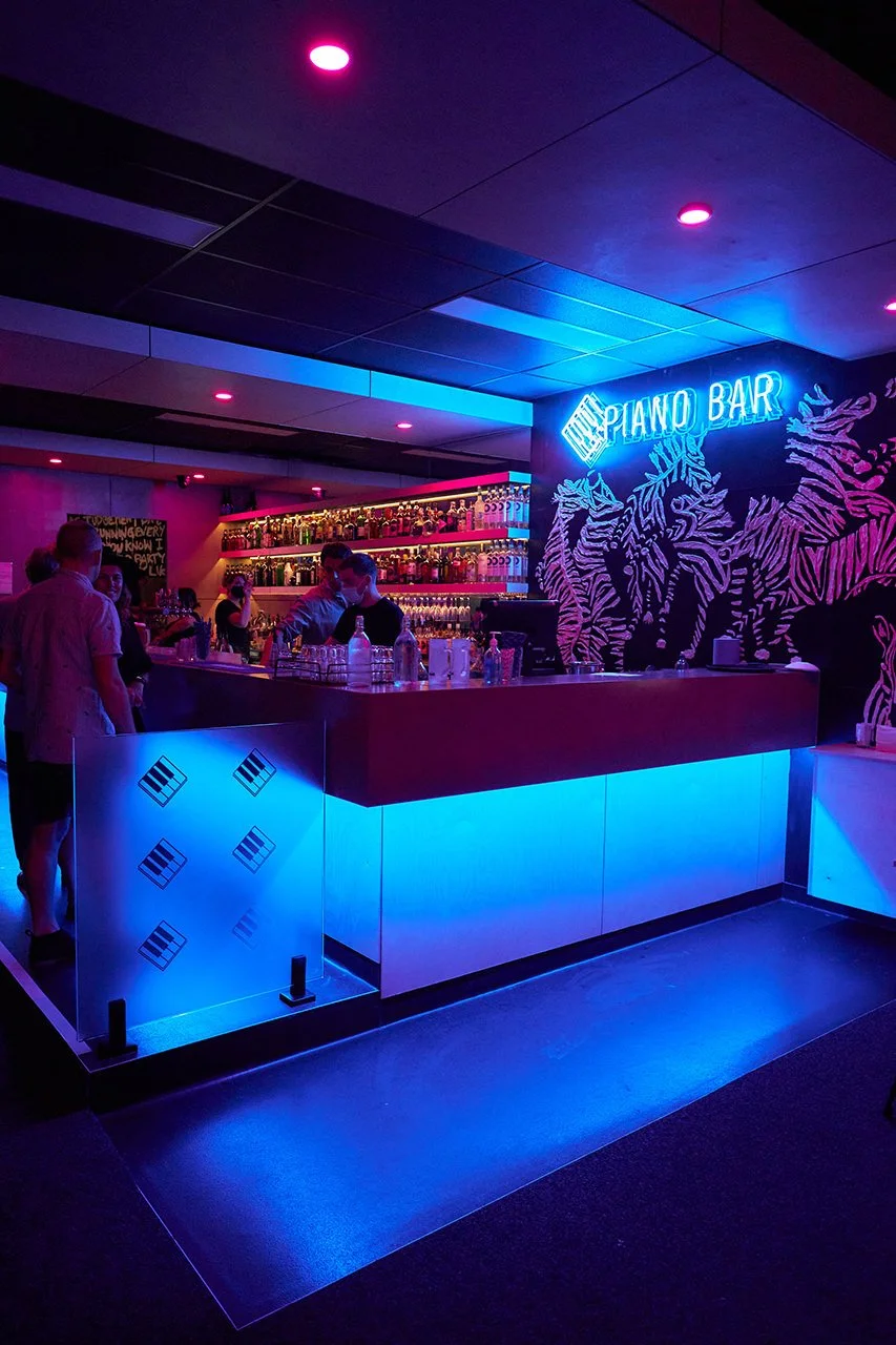 Neon-lit piano bar with a vibrant blue and pink lighting, bartenders preparing drinks behind a bar, and a wall mural of plants with a neon sign that reads 'Piano Bar'.
