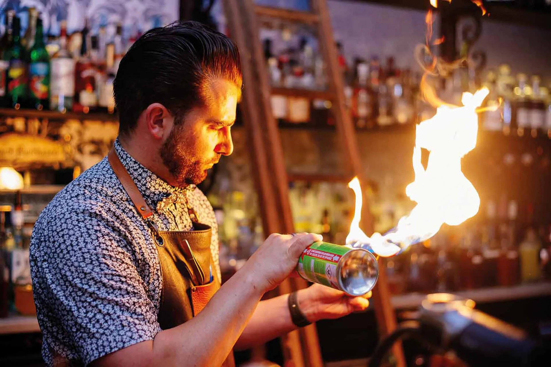 A bartender with dark hair and a patterned shirt, wearing an apron, is creating a flame with a fireproof spray can in a bar with liquor bottles in the background.