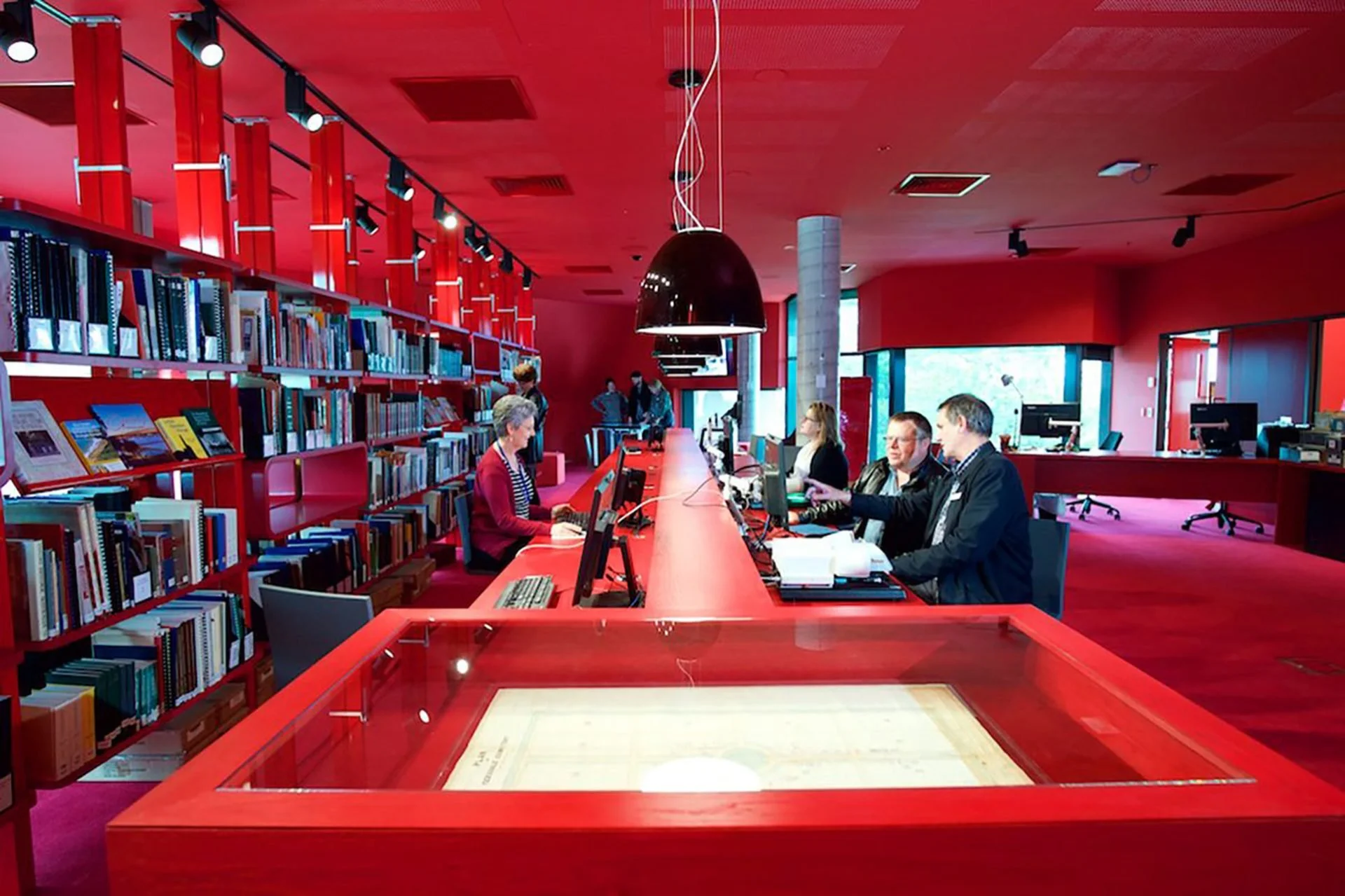 People working at a long red counter with computers in a red-walled library or information center.
