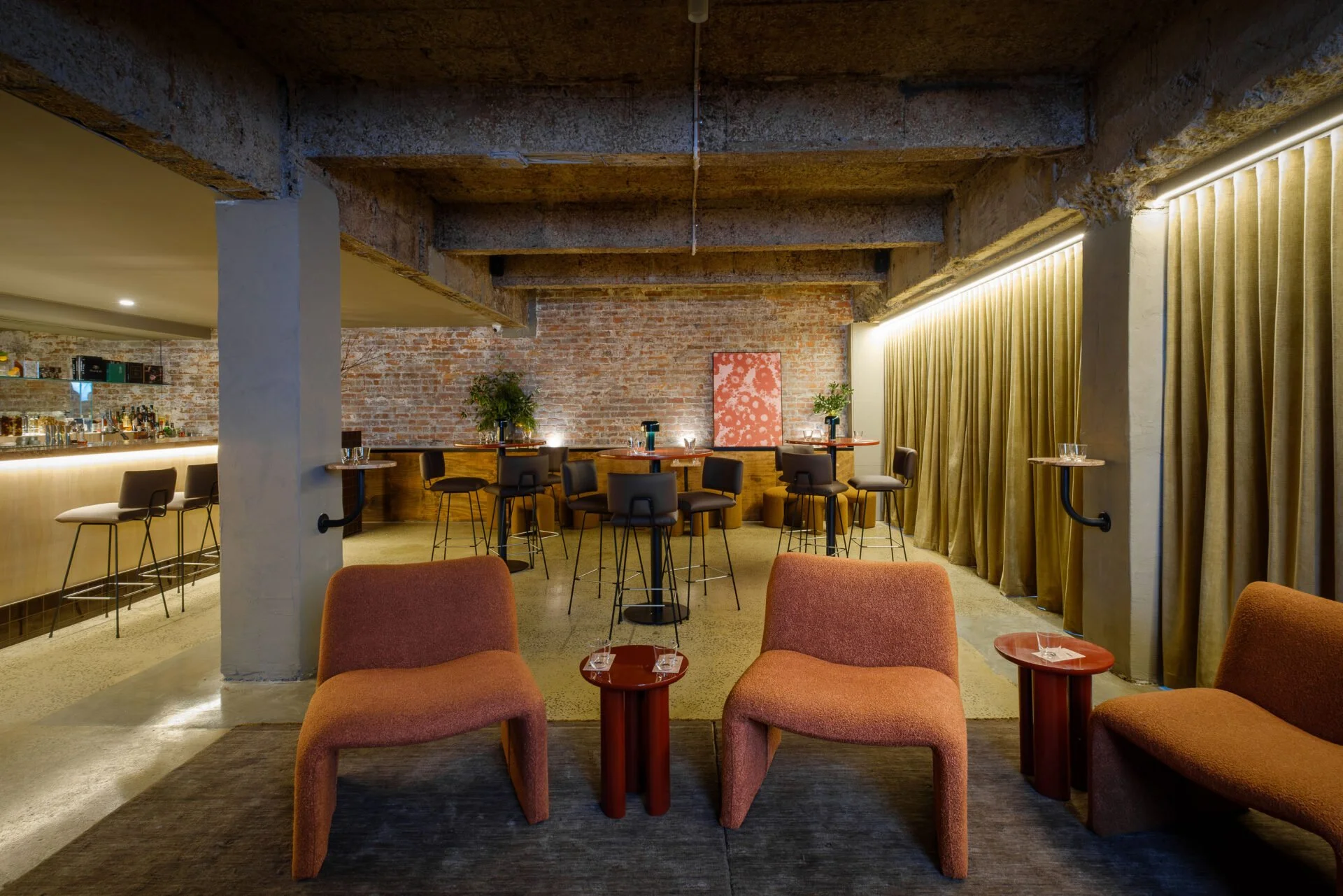 Interior of a modern bar or lounge with rust-colored chairs and small tables in the foreground, and a brick wall with a painting behind a high table and bar area in the background. Gold curtains run along one side of the room.