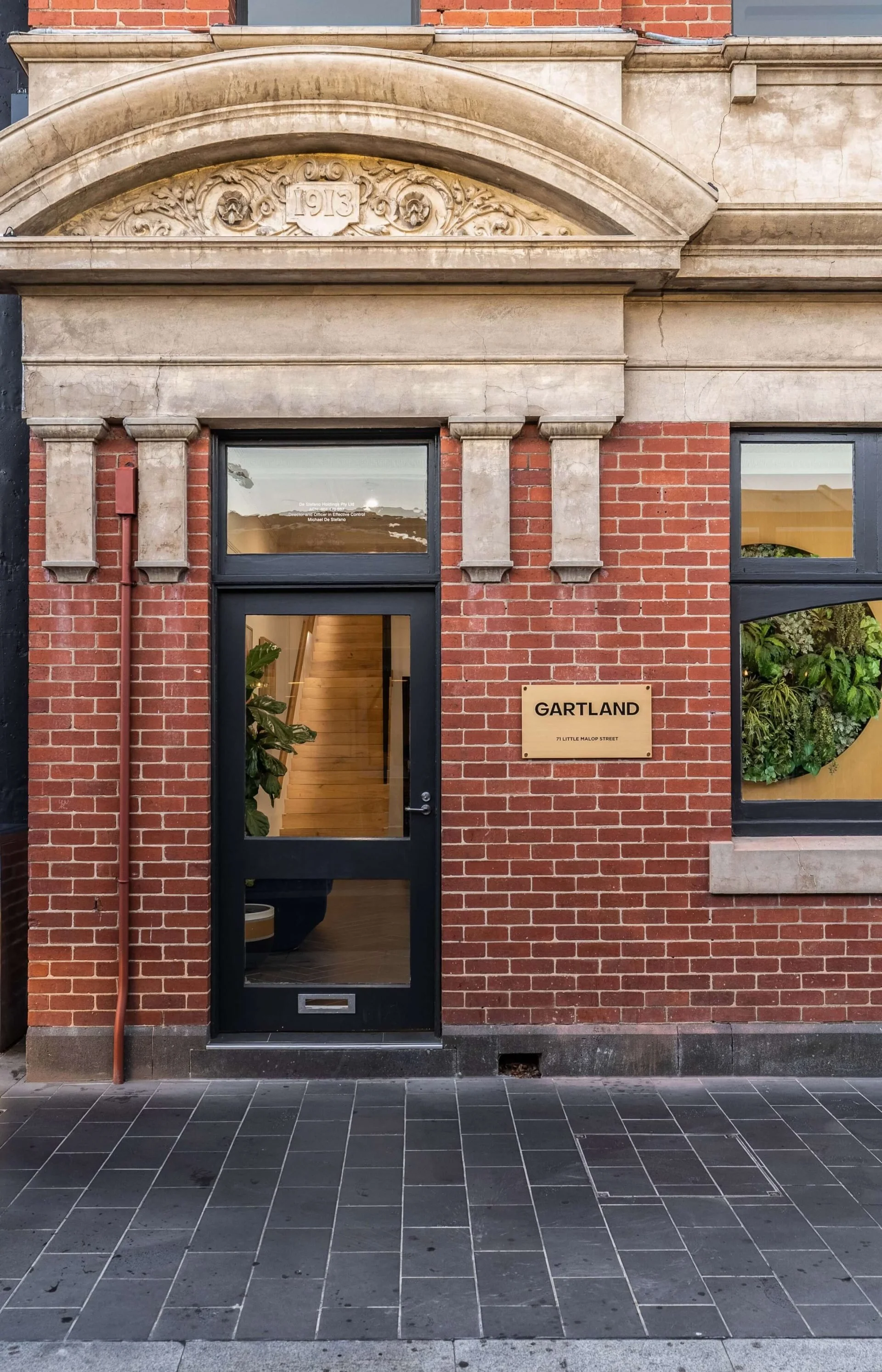 Brick building with stone decorative archway above the door, which has a window showing a staircase inside. There is a gold plaque on the wall reading "GARTLAND 71 LITTLE MALOP STREET."