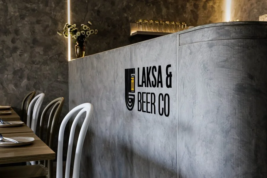 Interior of a restaurant with a concrete bar counter and white chairs. A logo on the bar reads 'Laska & Beer Co' with a stylized beer mug icon. There are flowers in a vase on a shelf and a decorative wall with warm lighting.