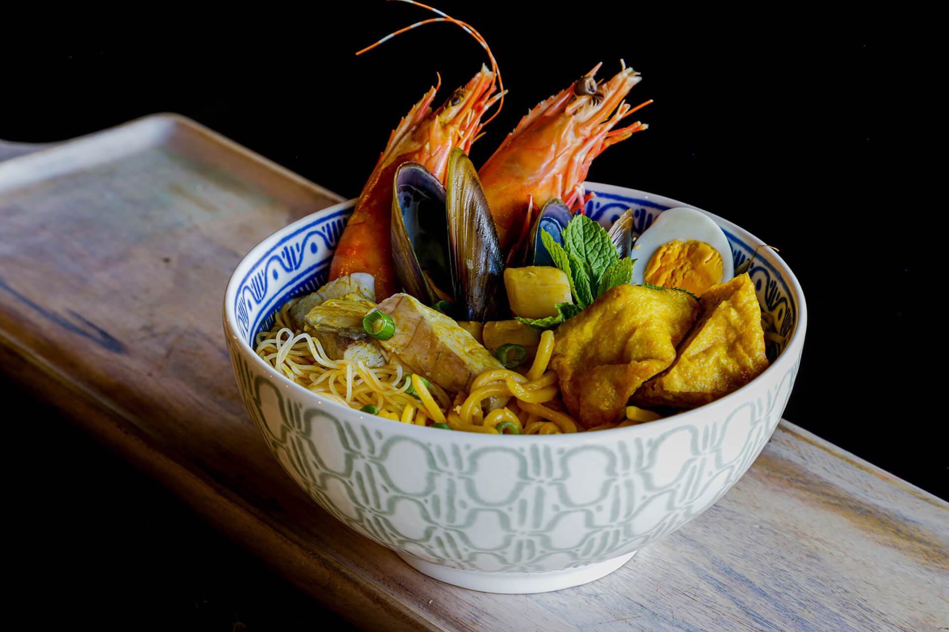 Bowl of Asian seafood noodle soup with shrimp, mussels, egg, chicken, and herbs on a wooden board.