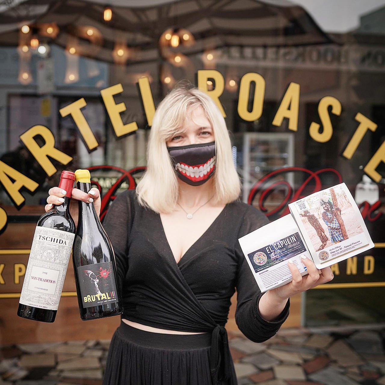 A woman with blonde hair wearing a face mask with a big smiling mouth print, holding two bottles of wine in one hand and a flyer and brochure in the other, standing outside a restaurant with a large window and yellow lettering.