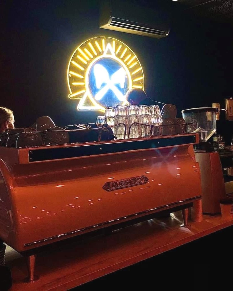 A neon sign of a coffee cup with a sun behind it on a dark wall, above an orange vintage espresso machine with cups and glasses on top, in a dimly lit cafe or bar.