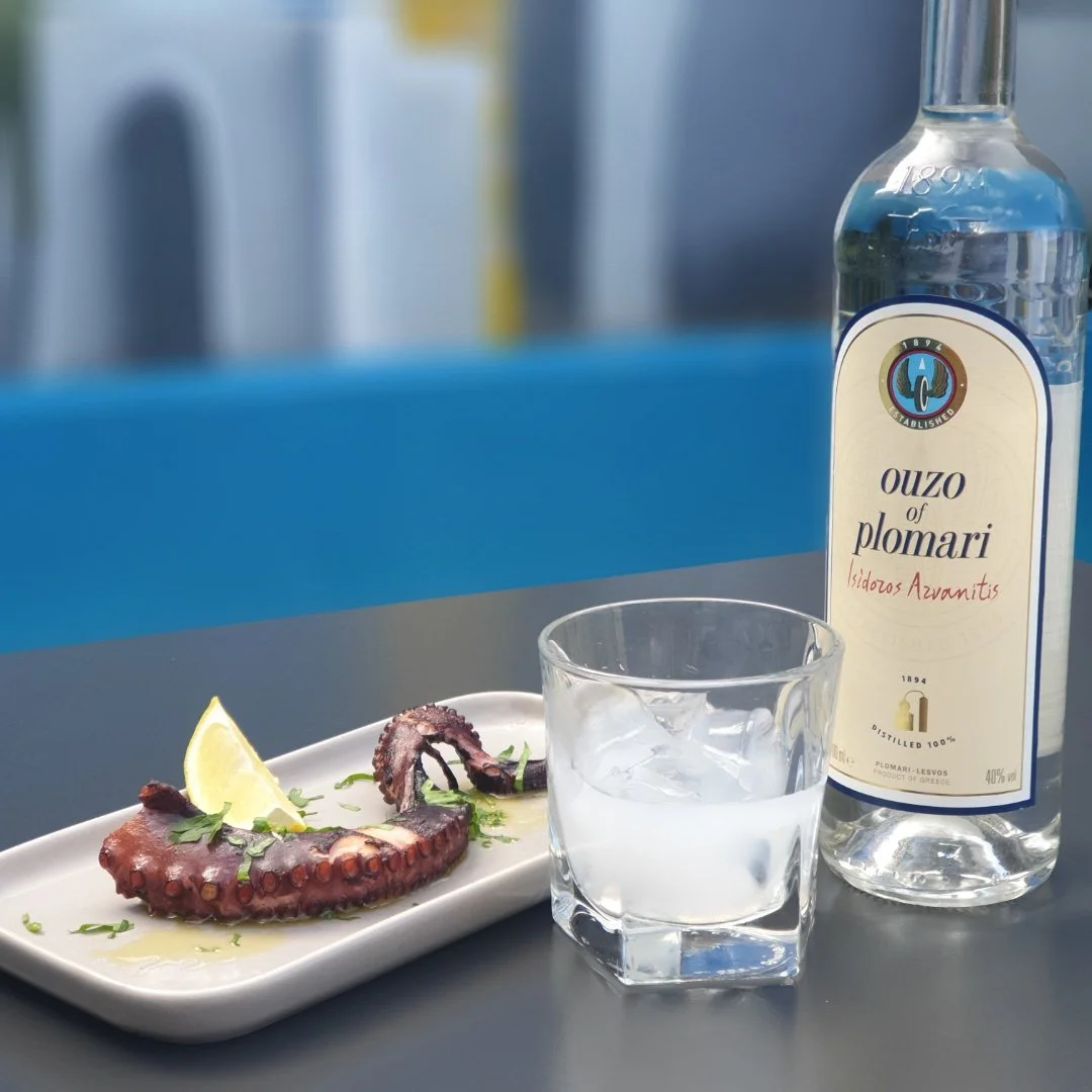 A plate with grilled octopus tentacle garnished with herbs and a lemon wedge, a glass of ice water, a bottle of ouzo, and a background with a blue wall.