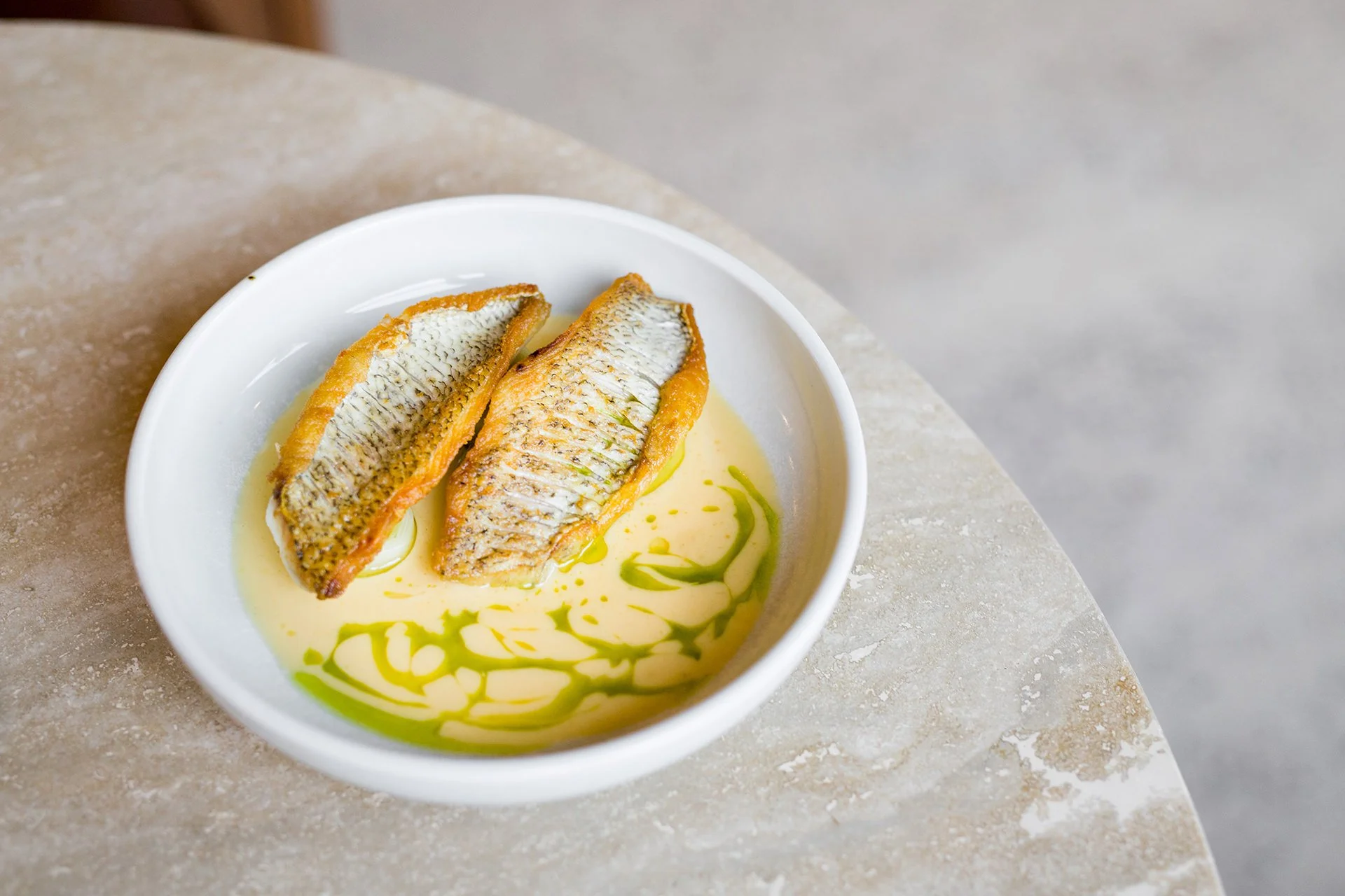 Two cooked fish fillets served on a white plate with a yellow sauce and green oil garnish