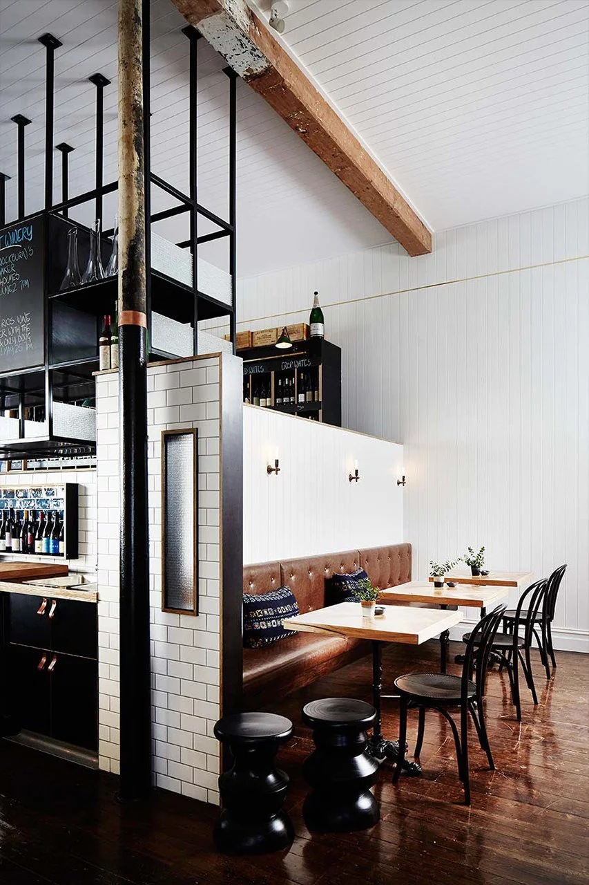 Interior of a modern restaurant with white paneled walls, wooden flooring, a brown leather banquette, small tables with white tops, and black chairs. Potted plants are on the tables, and decorative bottles are on a shelf above the white wall. A black