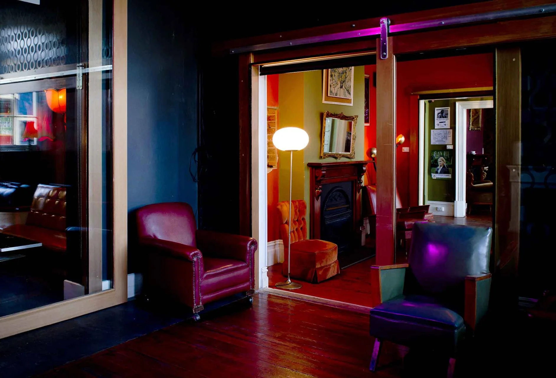 Interior of a cozy room with colorful walls, vintage furniture including a red leather armchair, an orange velvet chair, a green leather armchair, a standing lamp, a fireplace, and framed artwork on the walls.