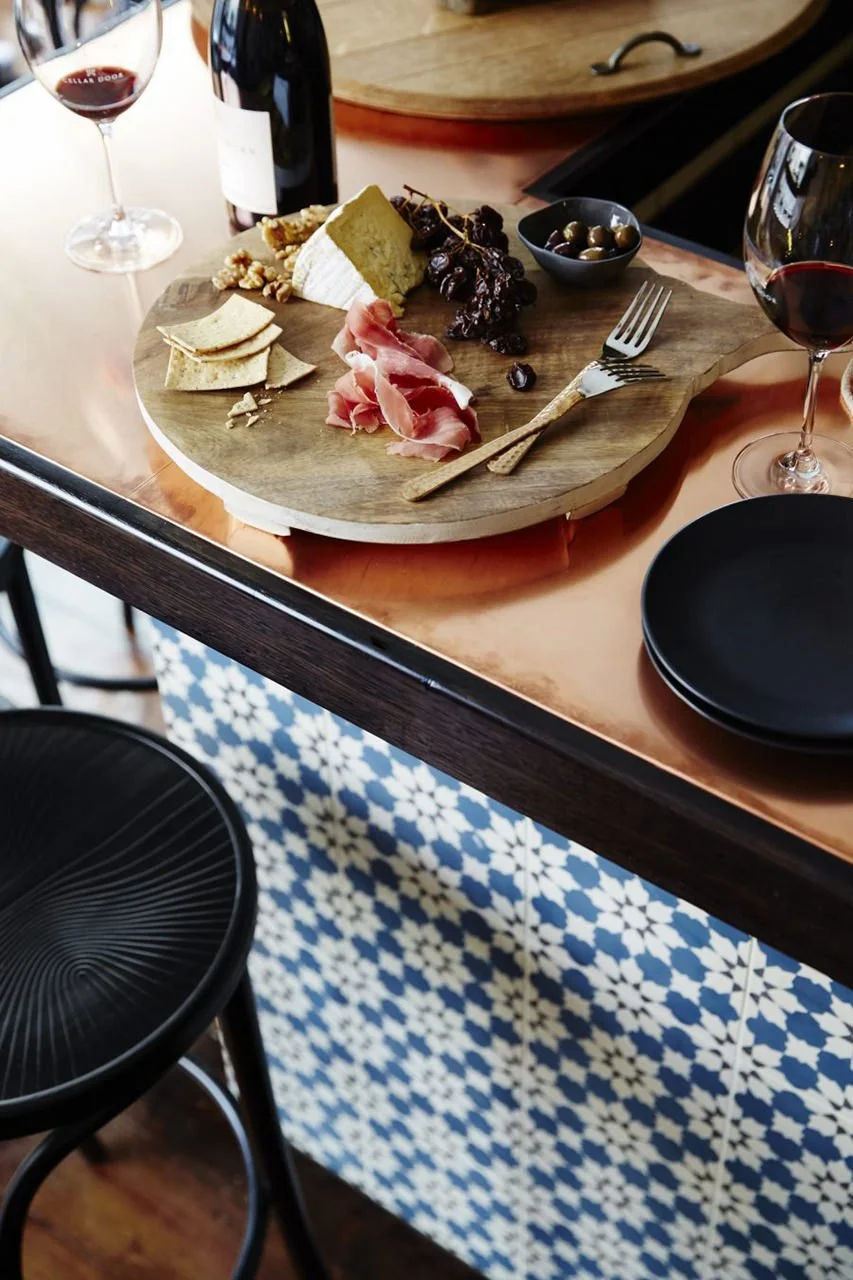A cheese and charcuterie board with grapes, dried fruit, crackers, and cheese, along with two glasses of red wine on a wooden table with black plates and a patterned tiled wall underneath.