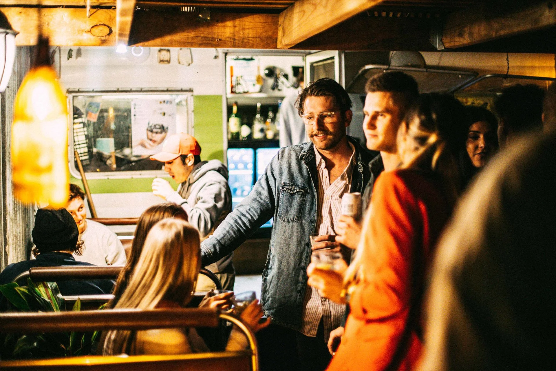 People socializing and drinking in a cozy bar with warm lighting, wooden decor, and a green wall in the background.