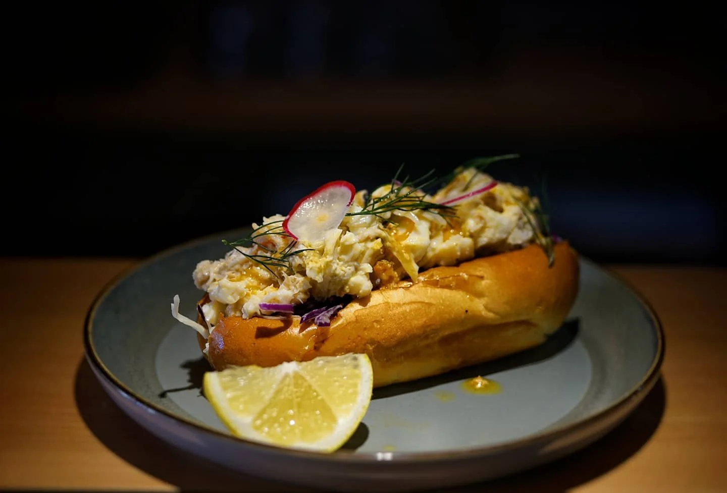 Lobster roll with chopped lobster meat, herbs, and vegetables on a bun, served with a lemon wedge on a plate.