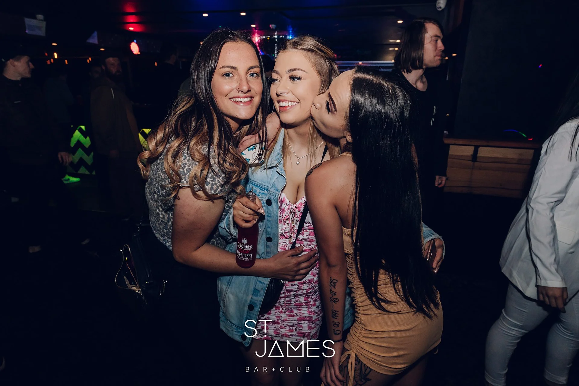 Three women smiling and posing closely together at a nightclub, with one woman kissing another on the cheek, in a dimly lit environment with other people in the background.