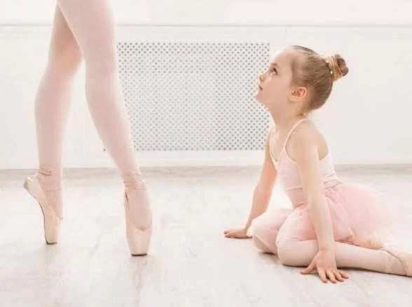 A young girl dressed as a ballerina sitting on the floor while looking up at an adult's legs in ballet shoes.