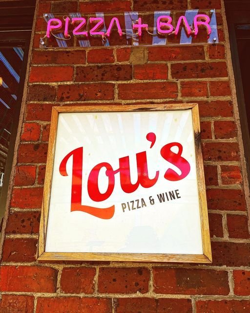 Sign for Loui's Pizza & Wine with a smaller neon sign above that says 'Pizza + Bar' in pink letters, mounted on a brick wall.