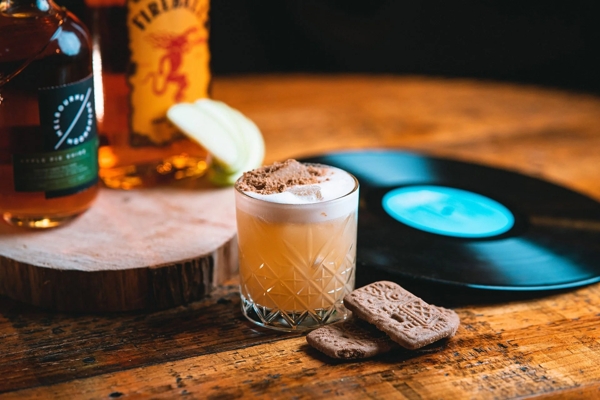 A whiskey sour cocktail garnished with a cinnamon stick and a small cookie, placed on a rustic wooden table with vinyl records and bottles in the background.