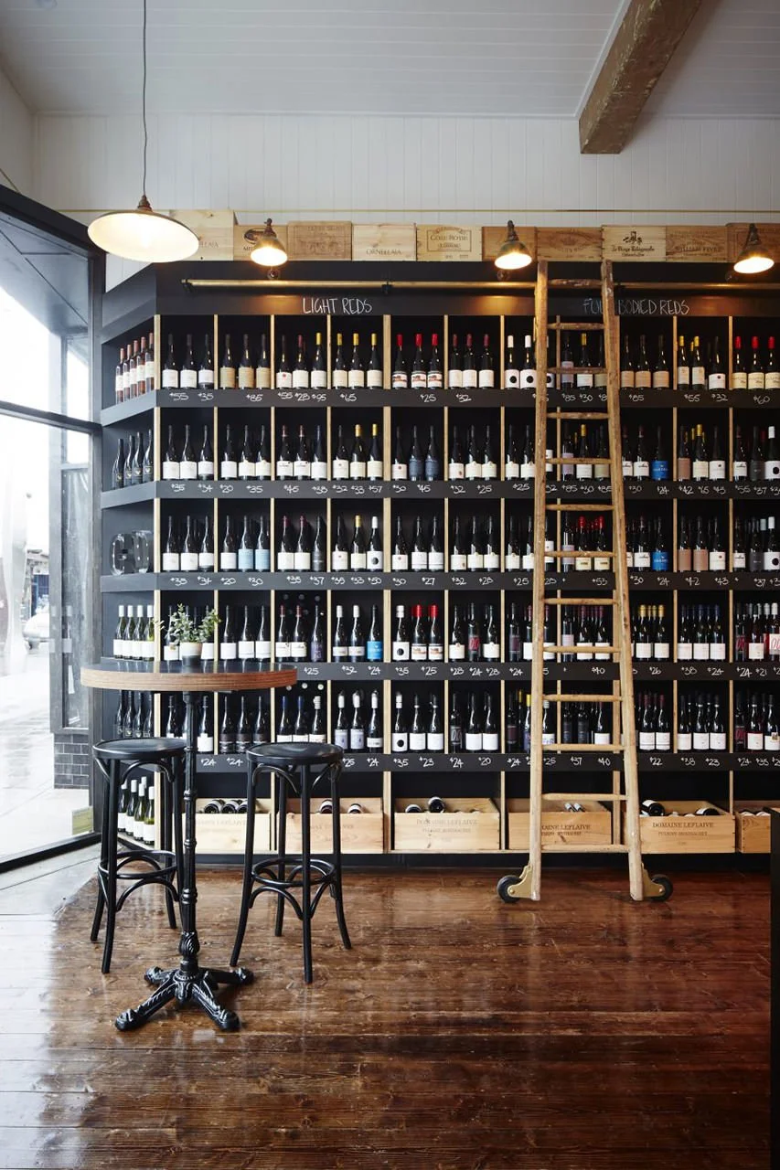 Wall of wine bottles displayed on a black shelf with a ladder, two black chairs, and a small round table in a wine shop or tasting room.