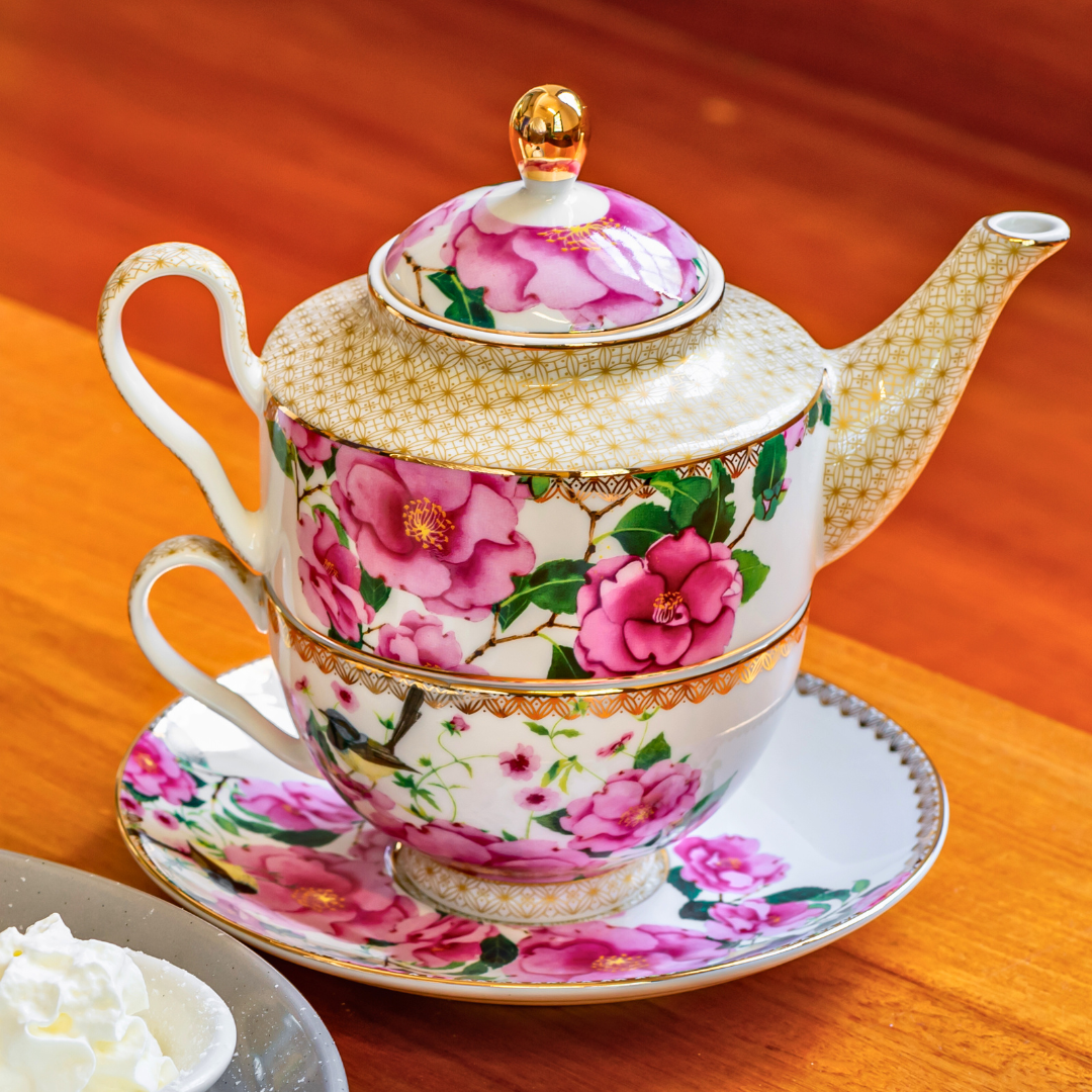 A decorative tea set with a floral pattern, consisting of a teapot, a cup, and a saucer, all adorned with pink flowers and gold accents, placed on a wooden table.