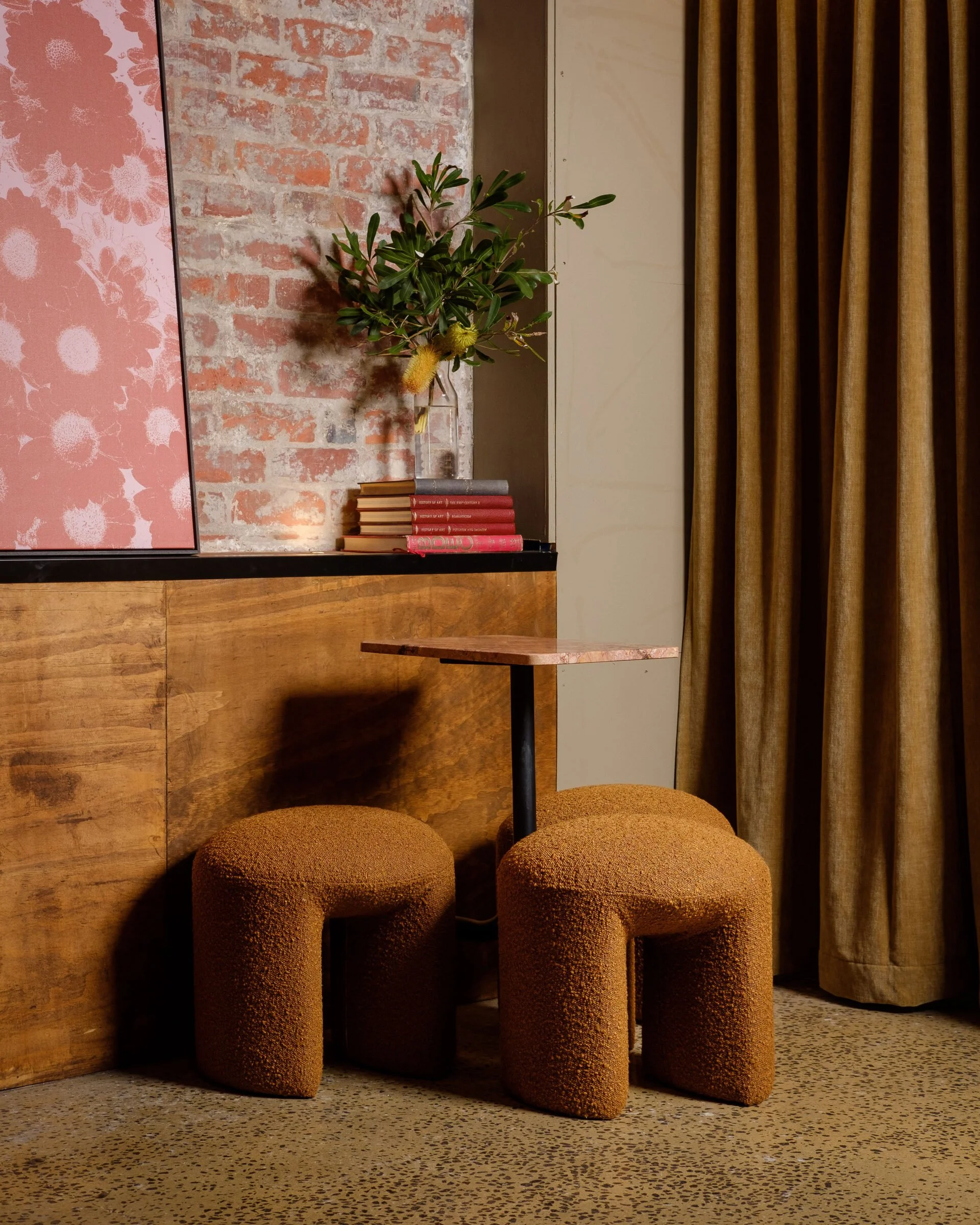Interior scene with a small round table and two textured orange stools, a wooden wall, a brick wall, a green plant in a vase, old books, a pink framed artwork, and beige curtains.