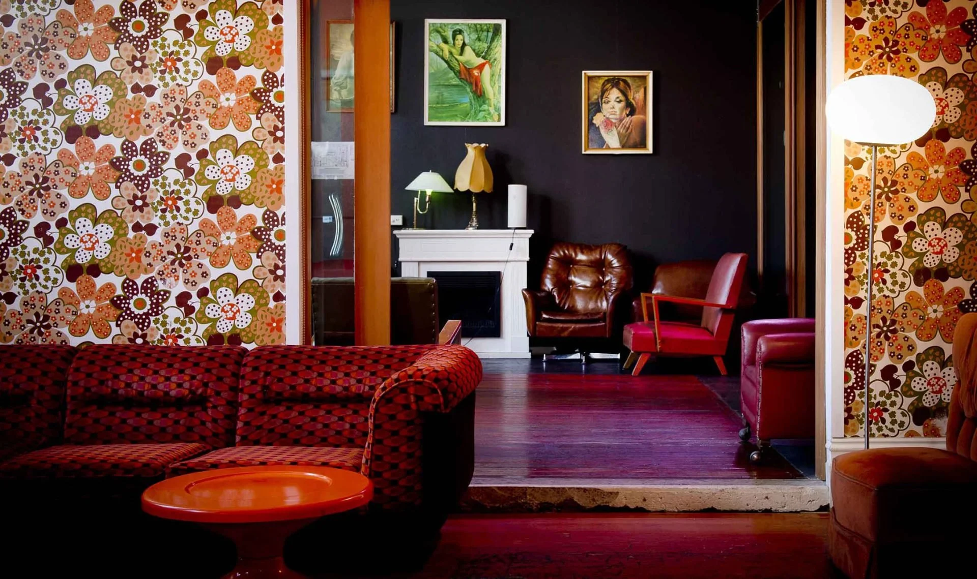 A cozy living room with patterned wallpaper featuring multicolored flowers, a red checkered sofa, and a round red table. There are various chairs, a fireplace with a white mantel, paintings on the wall, and a tall floor lamp with a white shade.